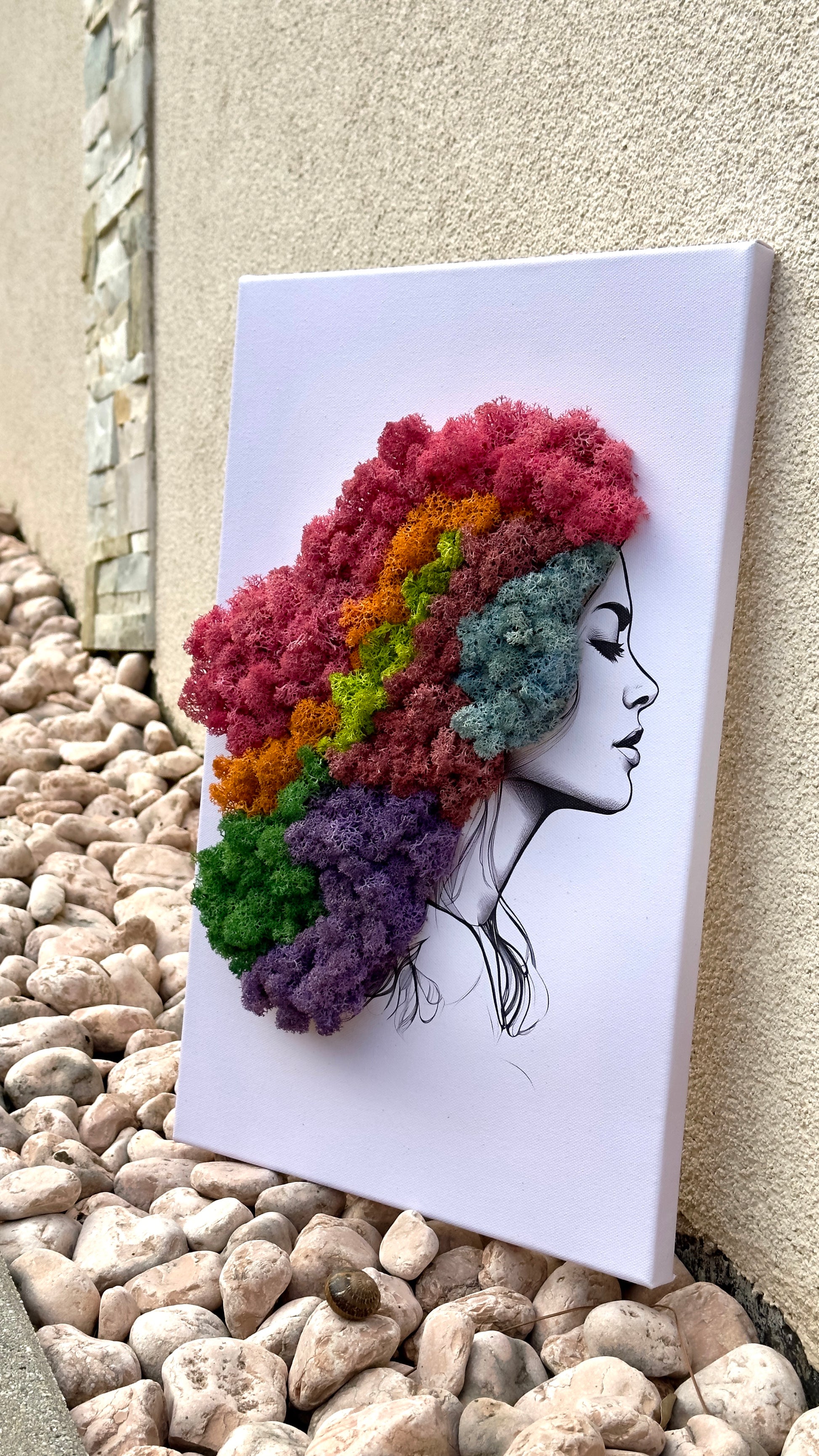 Canvas art of a woman with colorful preserved moss hair on a pebble surface.