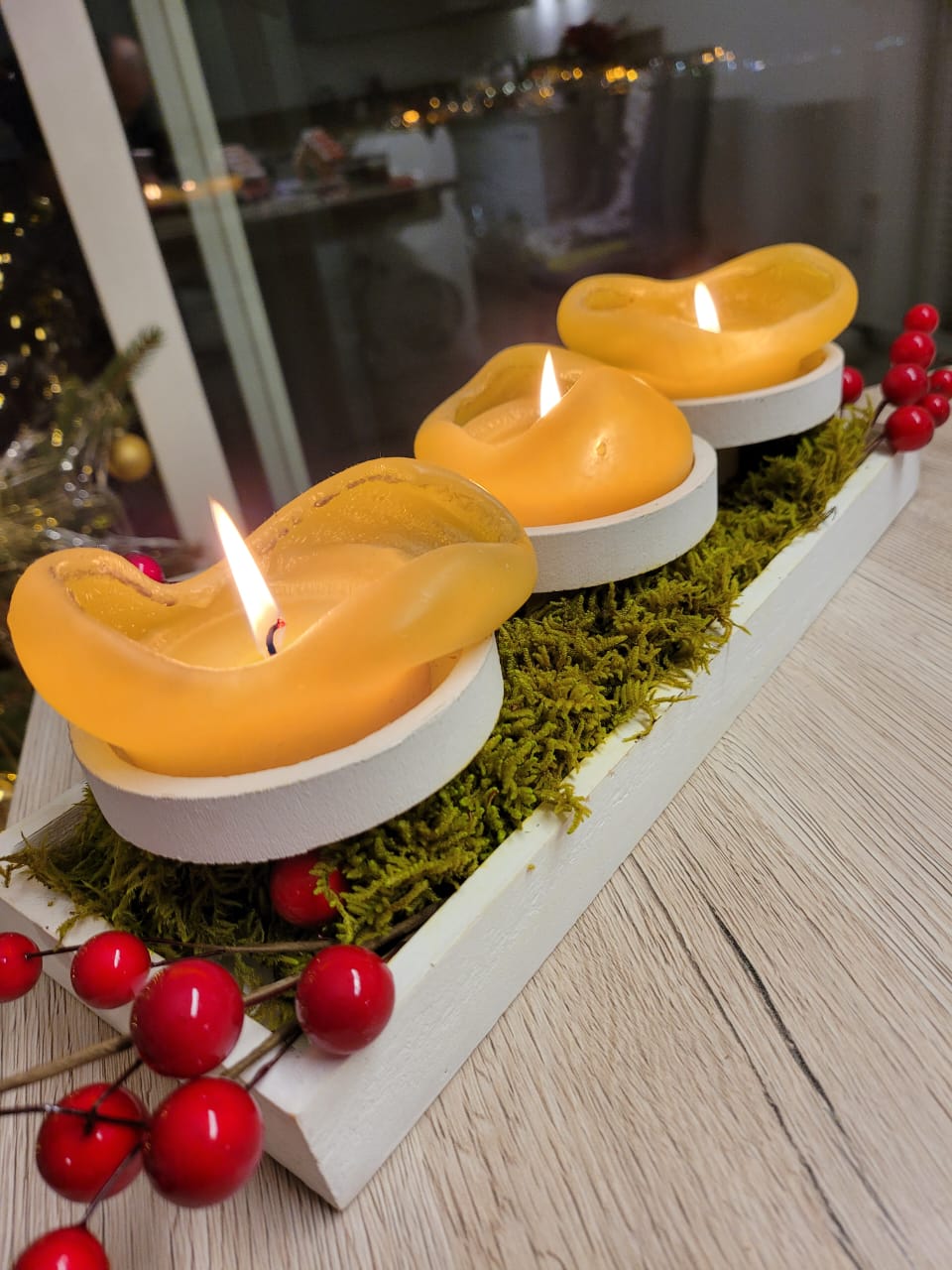Three yellow candles in white holders on a bed of moss with red berries on a wooden surface.