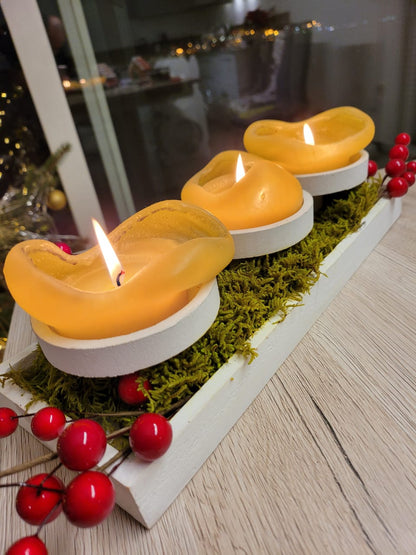 Three yellow candles in white holders on a bed of moss with red berries on a wooden surface.